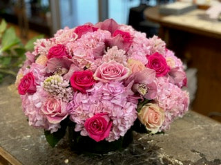 A floral arrangement featuring pink hydrangea, pink roses, hyacinth and orchid heads in a low cylinder vase. - Jerome Florists NYC