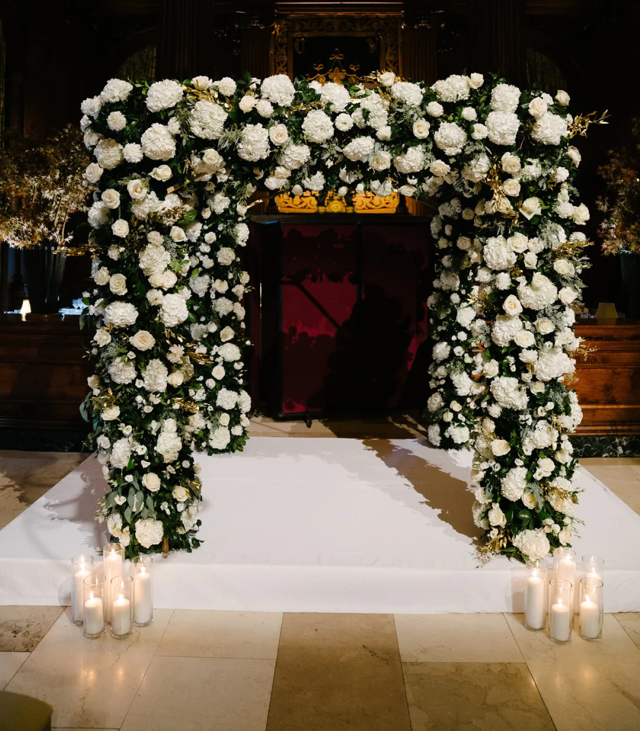 Floral archway with white flowers and greenery, adorned with candles on a white carpeted floor. Jerome Florists - NYC Since 1929