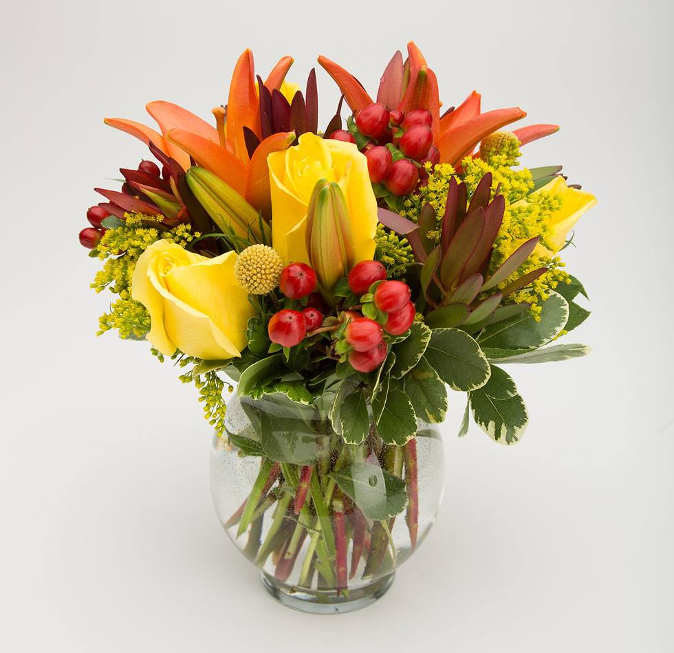 Arrangement of flowers with yellow roses, red berries, orange tulips and green leaves in a clear glass vase on a white background. - Jerome Florists NYC